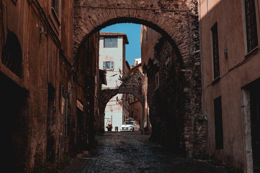 A narrow cobblestone street flanked by old buildings, an arched stone bridge, and a distant white building. 3