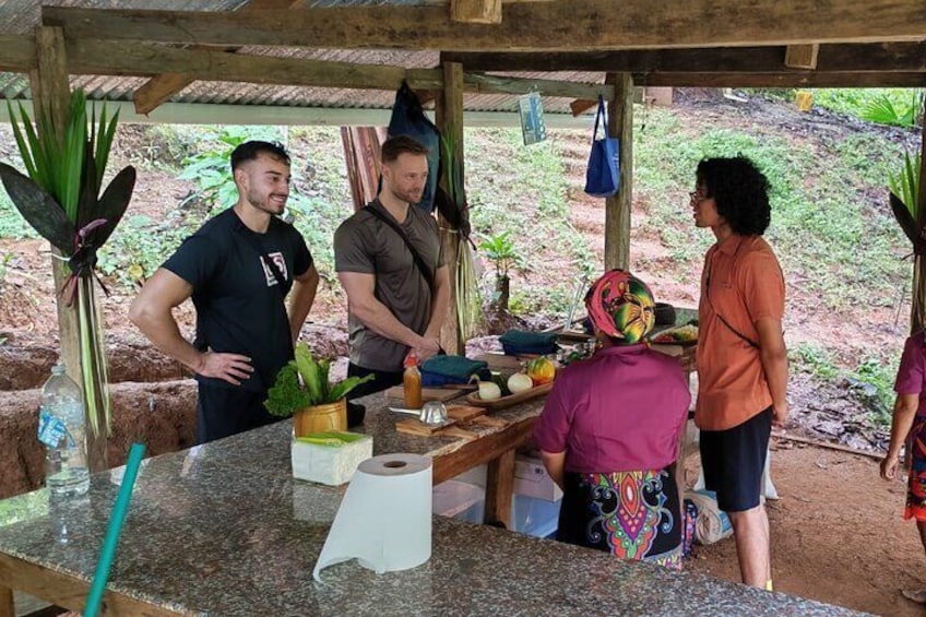 A group of people at an outdoor market with a wooden structure, various fruits and vegetables on display, and a person preparing food. 5