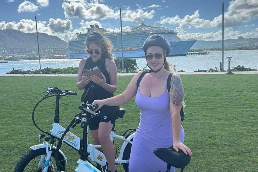 Two people on electric bikes, a grassy field, a body of water, and a distant building. 3