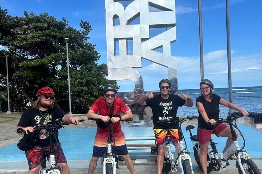 Four people on bicycles near a large sculpture and a body of water. 1