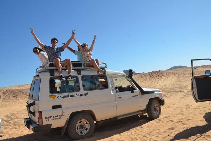 Un vehículo todoterreno blanco con tres personas encima, estacionado en un desierto con dunas de arena al fondo. 4