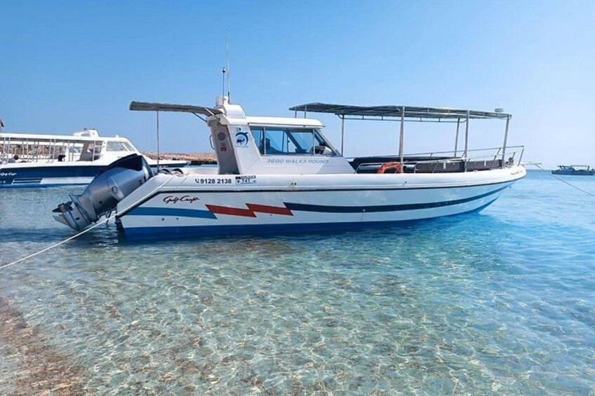 A small white boat with a blue stripe and red accents, anchored in clear shallow water with a clear sky in the background. 1