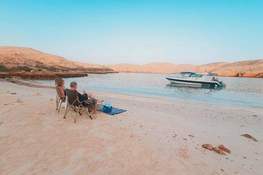 A sandy beach with two people sitting on chairs, a boat in the water, and a desert landscape in the background. 4