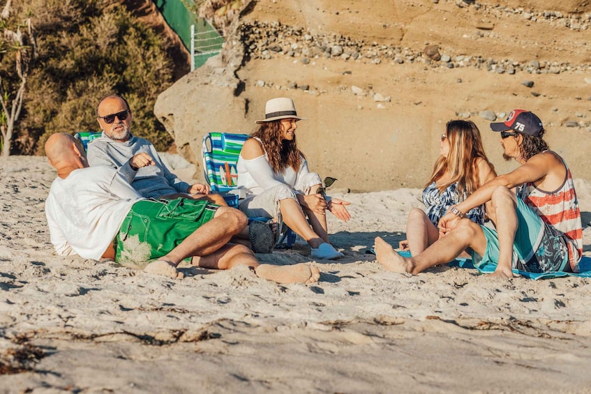 A group of people sitting on a sandy beach with a rocky cliff in the background. 3