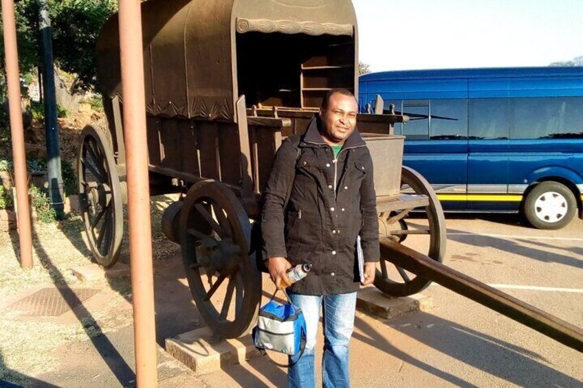 A man standing next to a wooden wagon with large wheels, holding a bag and a bottle. 1