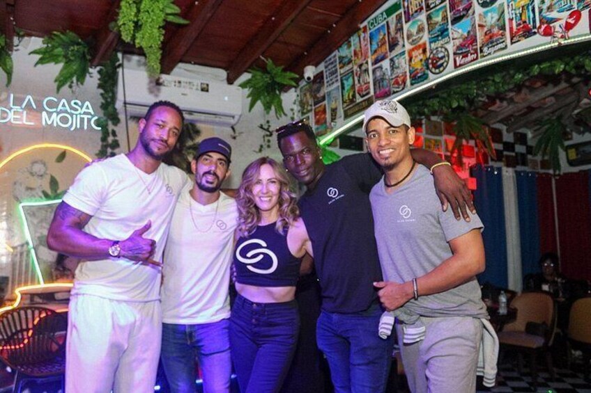 A group of people posing in a decorated indoor setting with a neon sign and tropical plants. 4