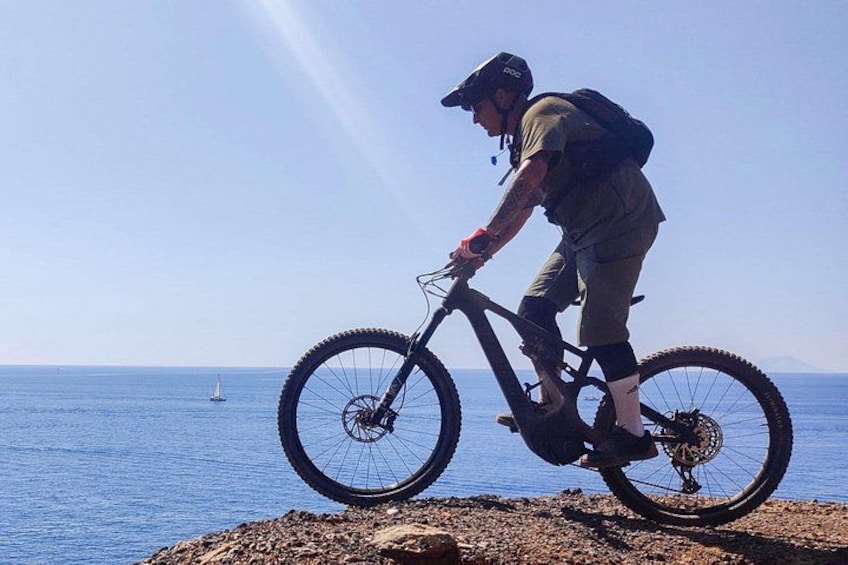 A person riding a mountain bike on a rocky shore with the ocean and a sailboat in the background. 4