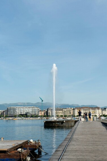 A waterfront with a wooden boardwalk, a fountain, boats, and buildings in the background. 3