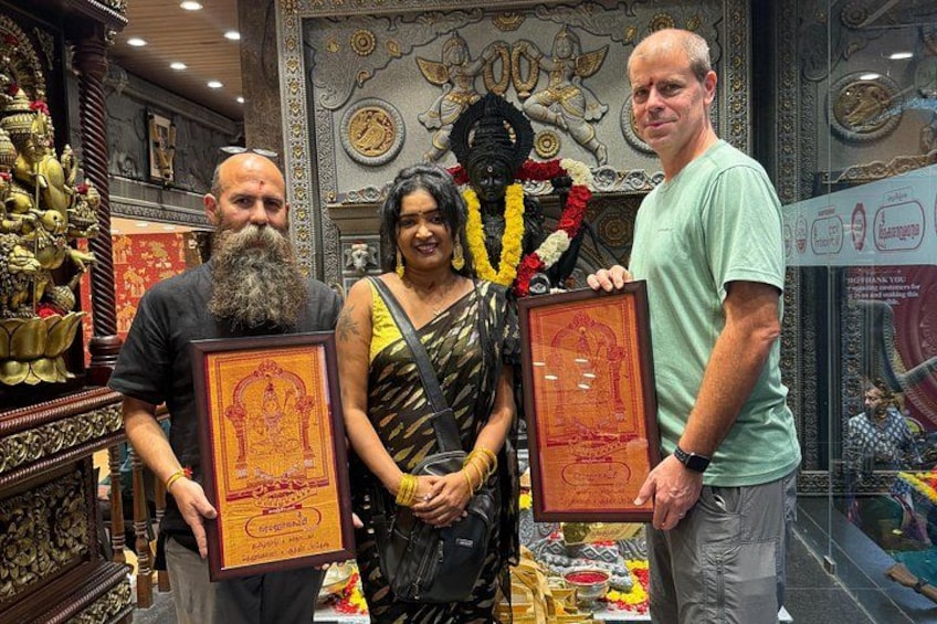 Three people holding framed certificates in a room with ornate decorations and a deity statue. 3