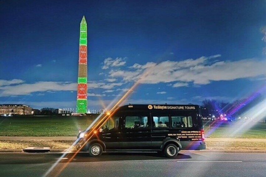 Een zwarte bus met de tekst 'Washington Signature Tours' rijdt over een weg, met een monument dat rood en groen is verlicht op de achtergrond. 1