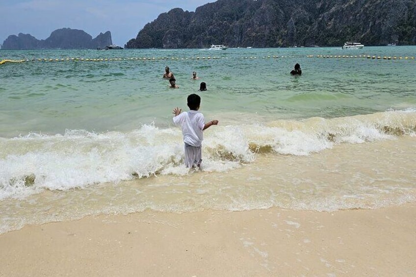 Un enfant joue dans une eau peu profonde sur une plage, où l’eau est d’un bleu turquoise clair et où l’on aperçoit, au loin, des îles rocheuses. 1