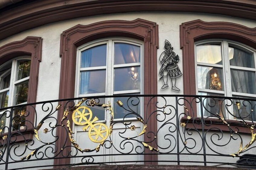 A building facade with decorative ironwork, a coat of arms, and a window with curtains. 5