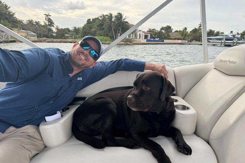 A man and a black dog on a boat with a water body and palm trees in the background. 5