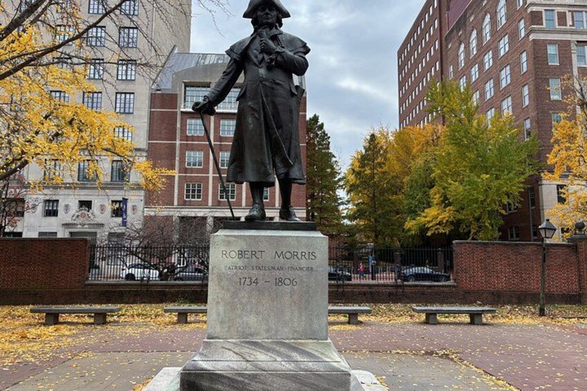 Een standbeeld van Robert Morris in een park met herfstbladeren, banken en gebouwen op de achtergrond. 5