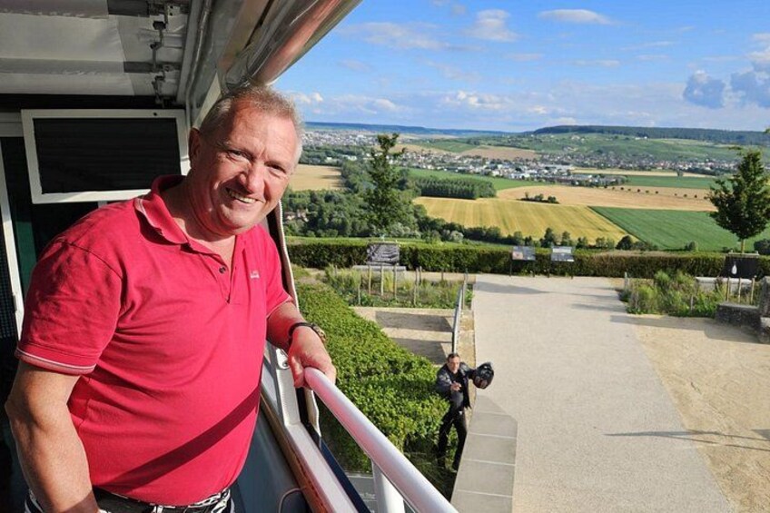 Un homme, vêtu d’un polo rouge, se tient sur un balcon d’où l’on peut admirer un paysage magnifique, avec des champs et une ville qui se dessinent à l’horizon. 2
