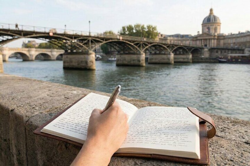 A hand writing in a book on a stone ledge with a river and bridges in the background. 3