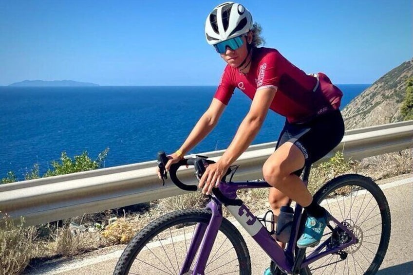 A cyclist riding a purple bike on a coastal road with a clear view of the sea and hills in the background. 5