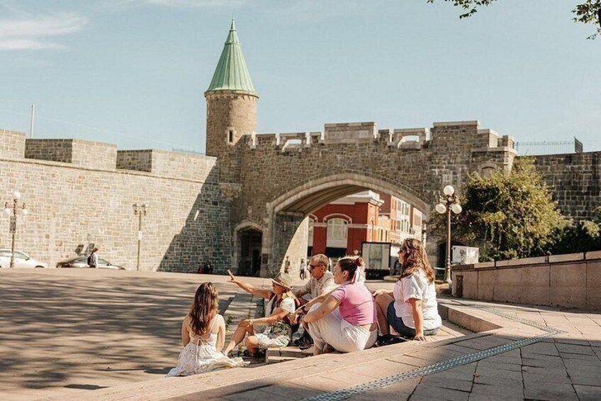A group of people sitting on a paved area in front of a historic stone wall with a tower. 4