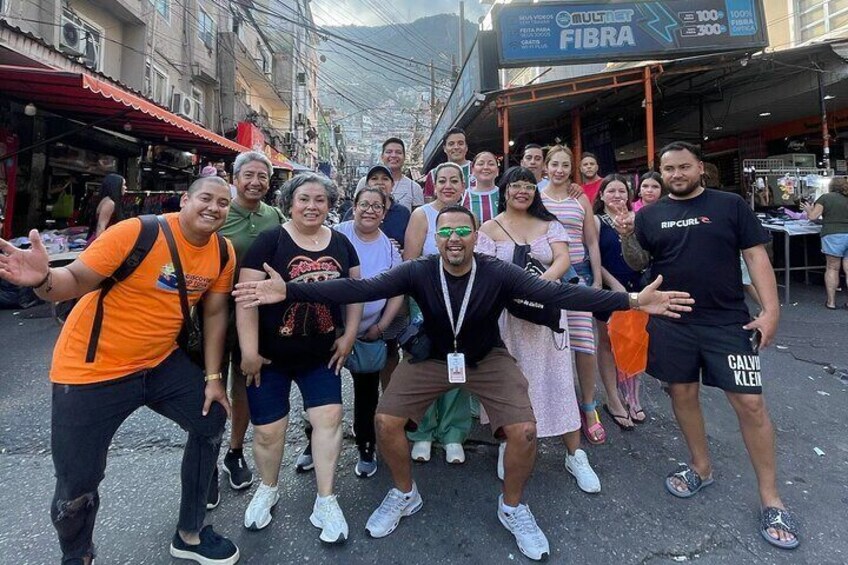Un groupe de personnes posant pour une photo dans une rue bordée de boutiques, avec une enseigne sur laquelle on peut lire « FIBRA ». 1