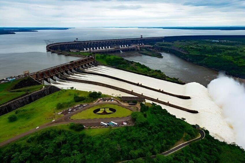 A large dam with water flowing over its spillway, surrounded by lush greenery and a body of water in the background. 2
