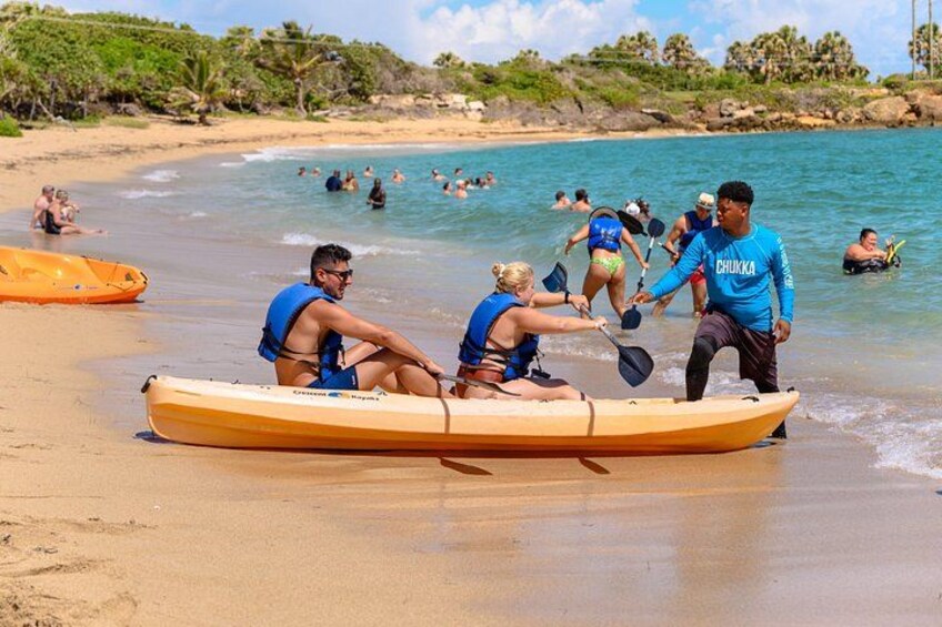 A group of people in a canoe on a sandy beach with water and palm trees in the background. 2