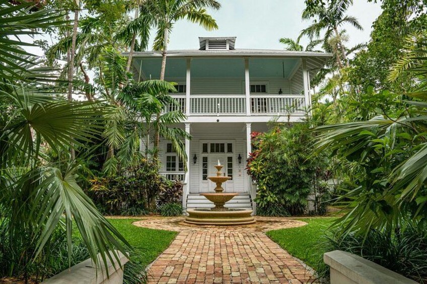 A white colonial-style house with a front porch, surrounded by lush tropical vegetation and a brick pathway leading to a central fountain. 4