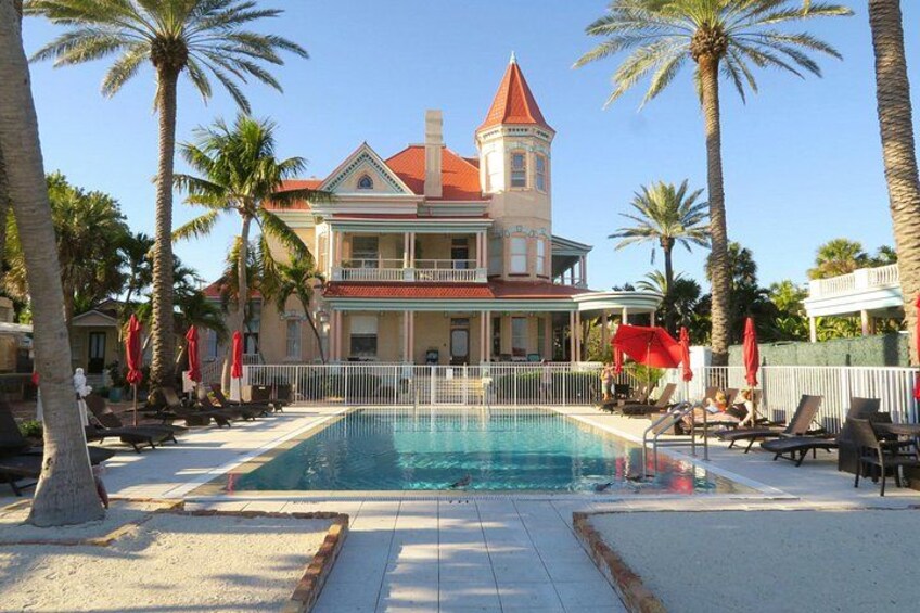 A swimming pool with lounge chairs and palm trees, a large building with a red roof in the background. 3