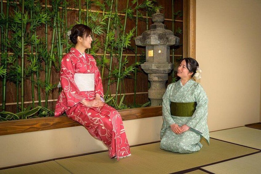 Two people in traditional Japanese attire sitting on tatami mats in a room with a bamboo backdrop and a stone lantern. 5