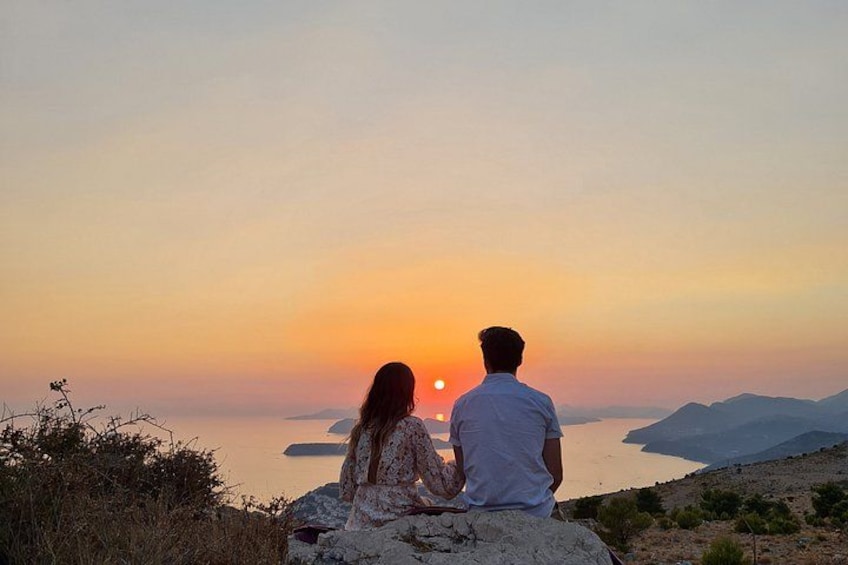 Zwei Personen sitzen auf einem Felsen und blicken auf einen Sonnenuntergang über dem Wasser, im Hintergrund Berge. 5