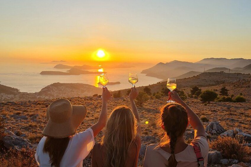 Drei Personen mit Weingläsern, die einen Sonnenuntergang über einer Küstenlandschaft mit Hügeln und einem Gewässer betrachten. 1
