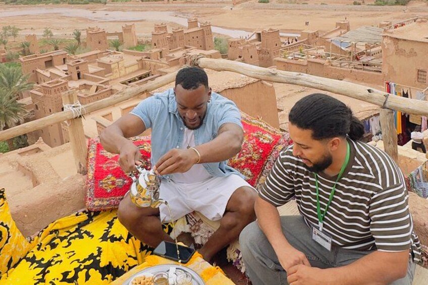 Deux personnes sont assises sur une plateforme en bois, un tissu rouge et jaune à motifs est étalé à leurs pieds, et elles sont entourées de bâtiments traditionnels construits en briques de terre. 4