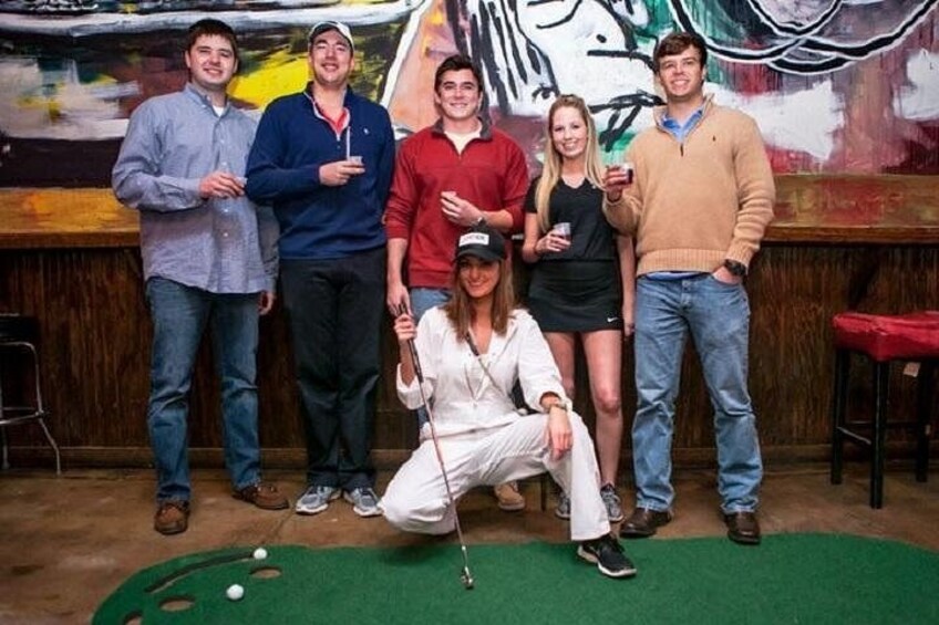 A group of people posing in a bar setting with a green felt surface and a white golf ball. 1