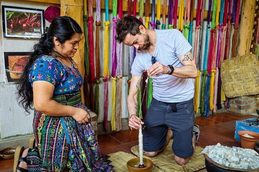Een persoon knielt op de vloer, bezig met een lang, dun voorwerp, mogelijk een gereedschap of instrument, in een kamer met kleurrijke hangende objecten op de achtergrond. 5