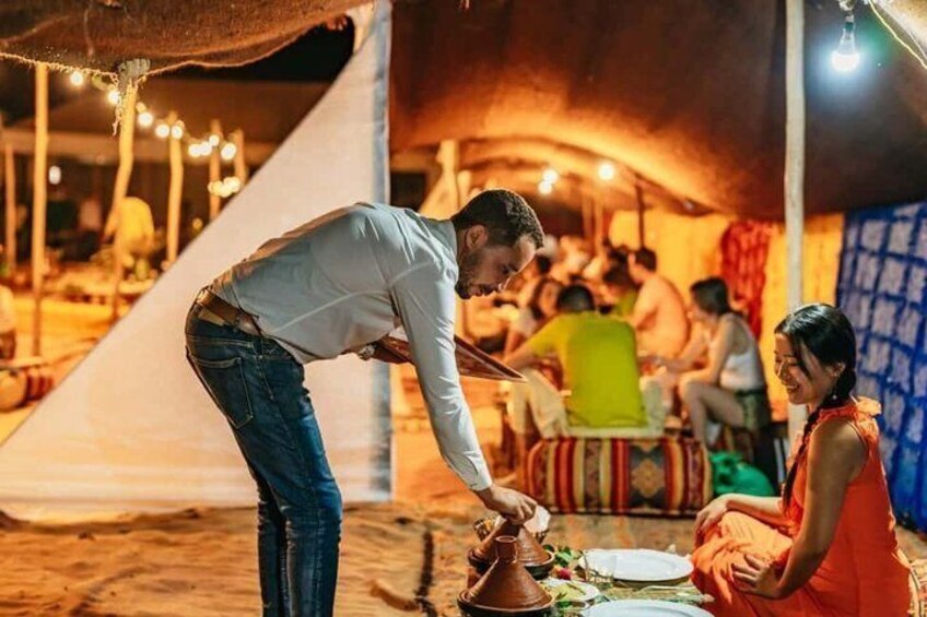 A person serving food in a tented area with people seated around a table. 1