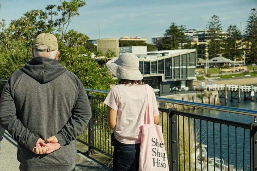 Two people standing on a bridge, overlooking a body of water with buildings and greenery in the background. 5