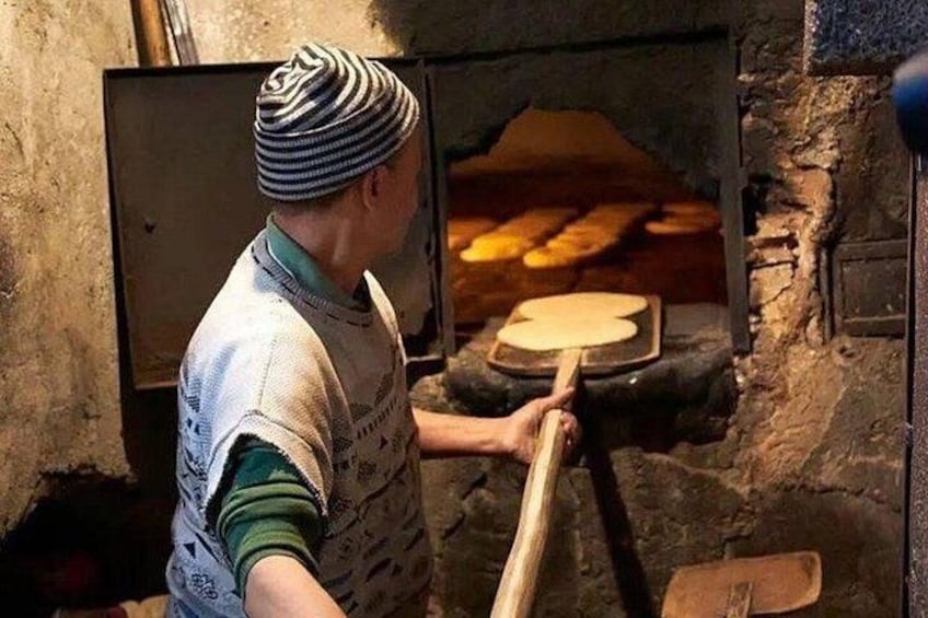 A person in traditional attire using a long-handled tool to manage a pizza in a brick oven. 3
