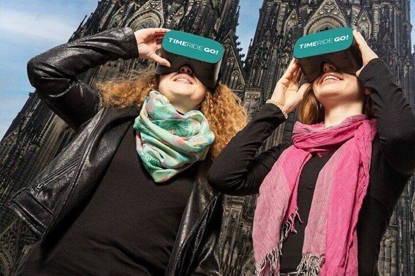 Zwei Personen mit VR-Brillen, auf denen 'TIMERIDE GO!' steht, vor einem Gebäude im neugotischen Stil. 1