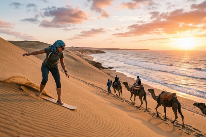 A person surfing on a sand dune, camels and riders in the background, and a sunset over the ocean. 2
