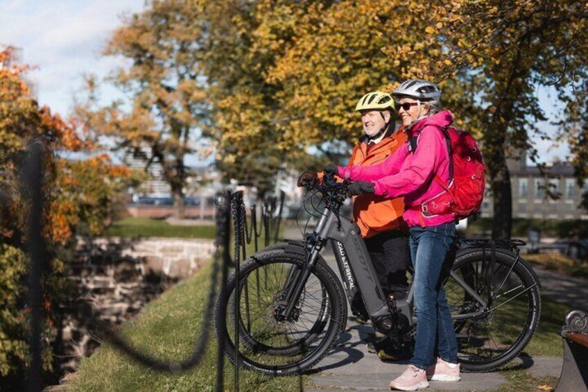 Zwei Personen stehen neben einem Fahrrad mit einer Ladestation, in einem Park mit Herbstbäumen. 2