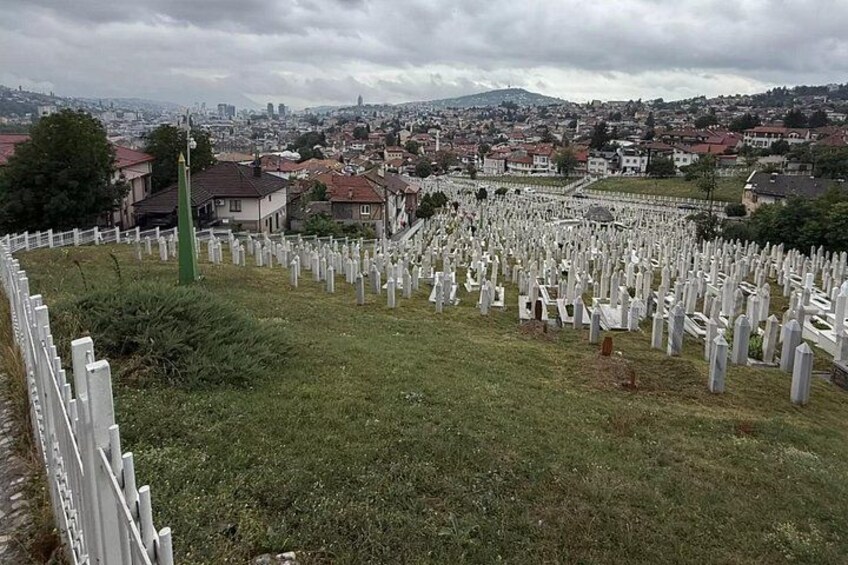 A cemetery with numerous white headstones, a green monument, and a cityscape in the background. 3