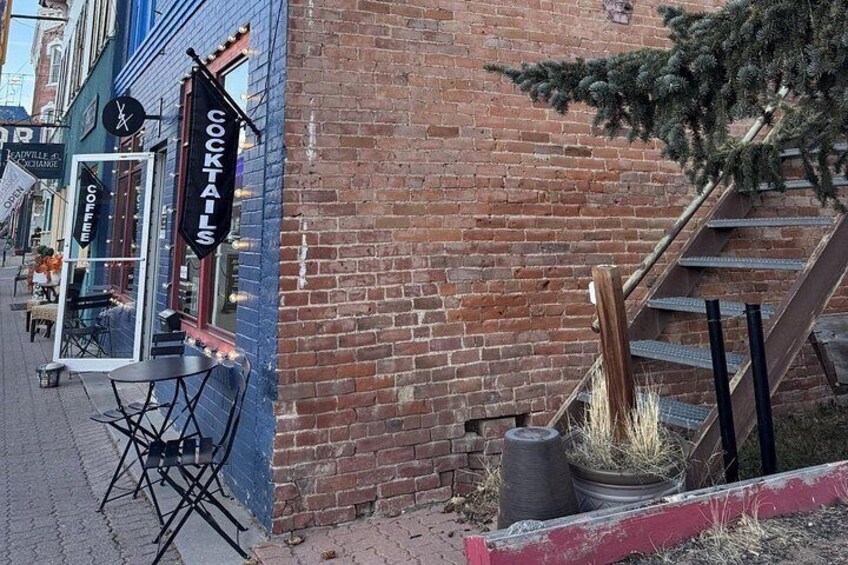Un edificio de ladrillo con un cartel anunciando cócteles y café. Hay mesas y sillas al aire libre, y una escalera que lleva a la entrada. 4