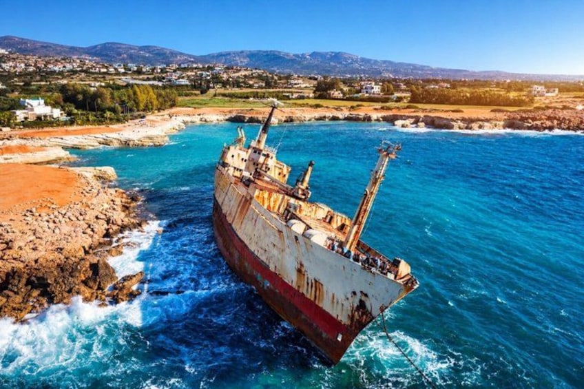 Een groot, roestig schip dat aan land is komen liggen op een rotsachtige kust, met helderblauw water en een verre horizon. 1