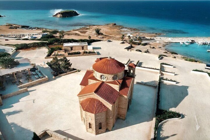 Une église côtière, avec un toit recouvert de tuiles rouges, entourée d’une zone sablonneuse et donnant sur la mer. 1
