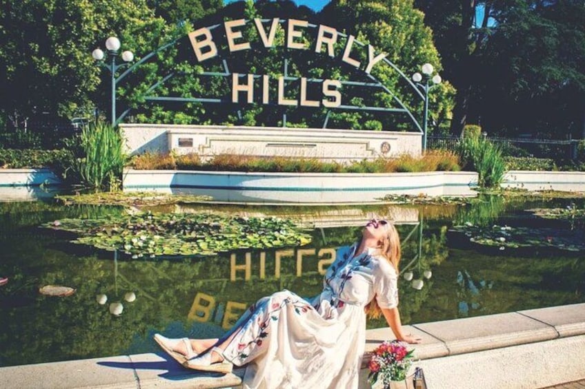 A woman in a white dress sitting by a pond with a sign reading 'BEVERLY HILLS' in the background. 5