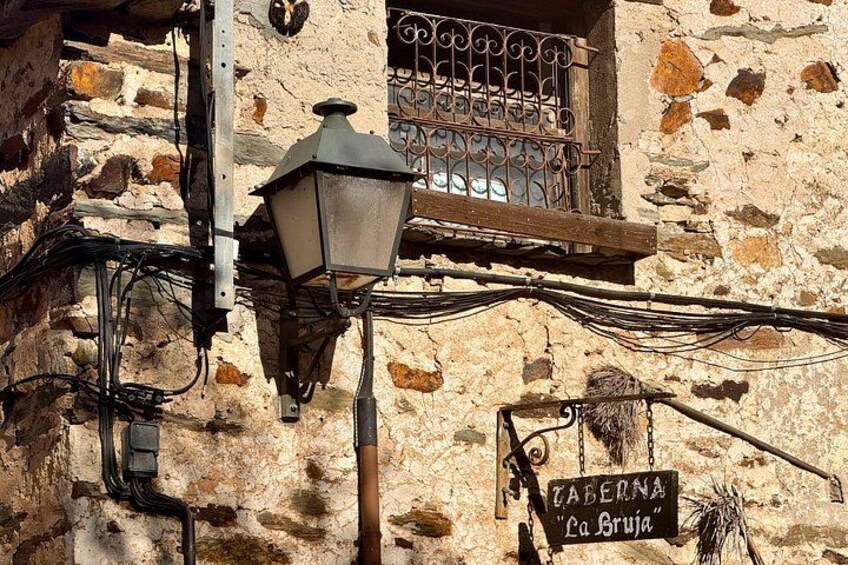 A rustic stone building with a metal window grille, a street lamp, and a sign reading 'CABERNA La Bruja'. 2