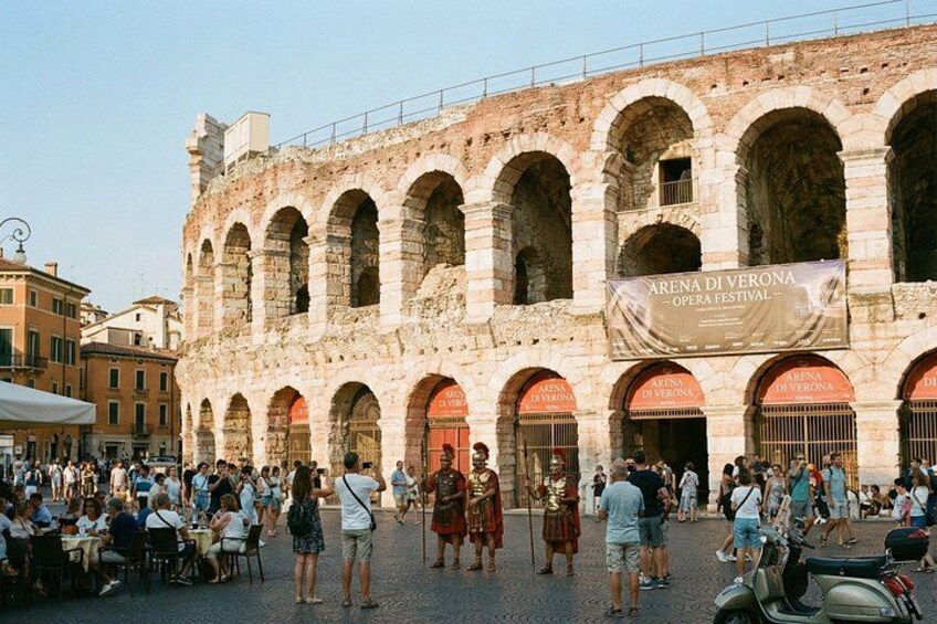 An outdoor area with a historic building featuring multiple arches and a banner for the Arena di Verona Opera Festival. 1