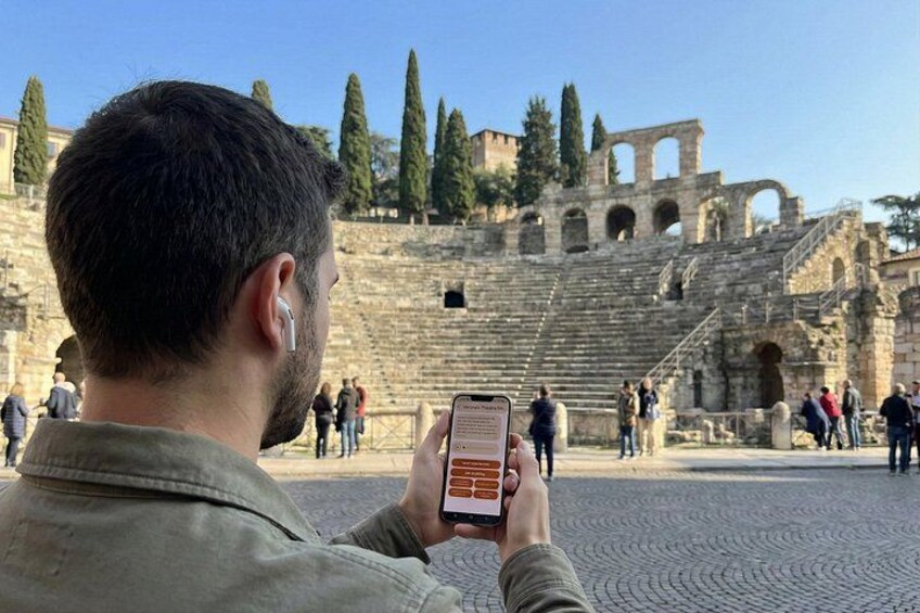 A person taking a photo of an ancient amphitheater with stone steps and columns. 3