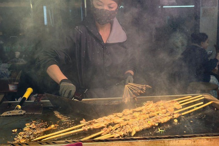 Jemand kocht auf einer großen Grillplatte, dazu liegen Spieße mit Essen, umgeben von Rauch. 5