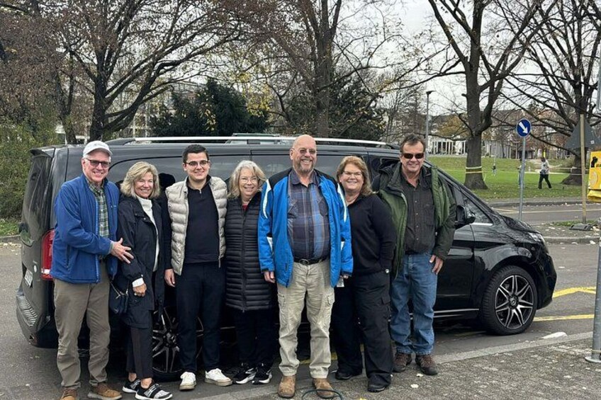 Un groupe de personnes se tient devant un fourgon noir, dans une rue bordée d’arbres, avec un autobus jaune en arrière-plan. 3