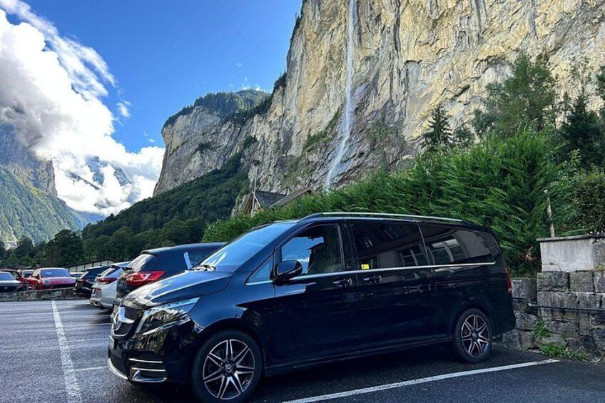 A black minivan parked in a parking lot with a mountainous backdrop and a waterfall. 3